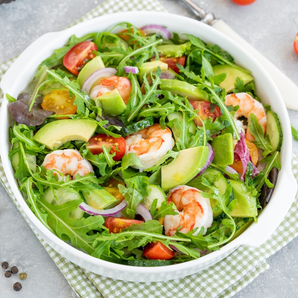 Shrimp and avocado salad with cherry tomatoes, red onion, and arugula in a white bowl.