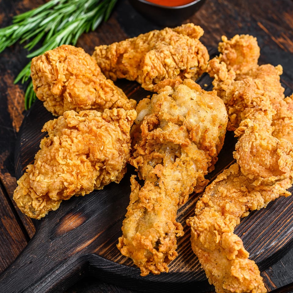 Crispy fried chicken pieces, including drumsticks and tenders, on a dark wooden board with rosemary.