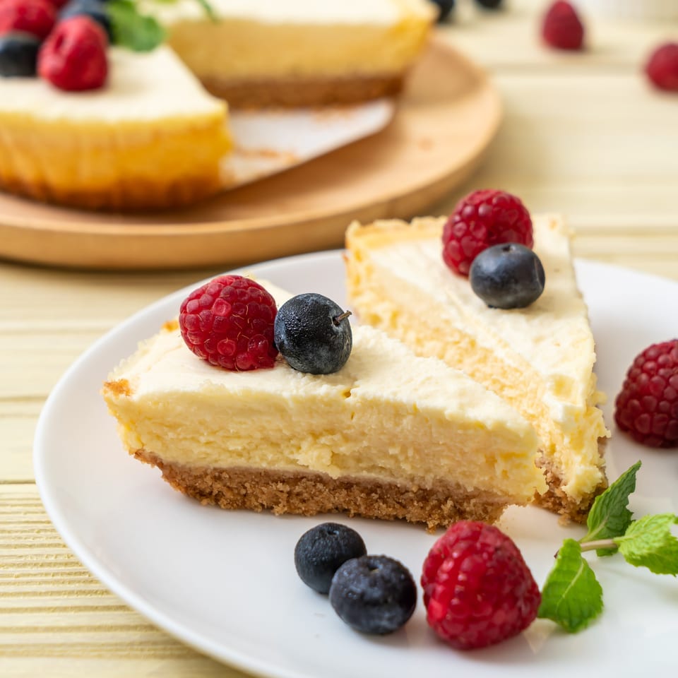 Two slices of cheesecake with raspberries, blueberries, and mint on white plates.